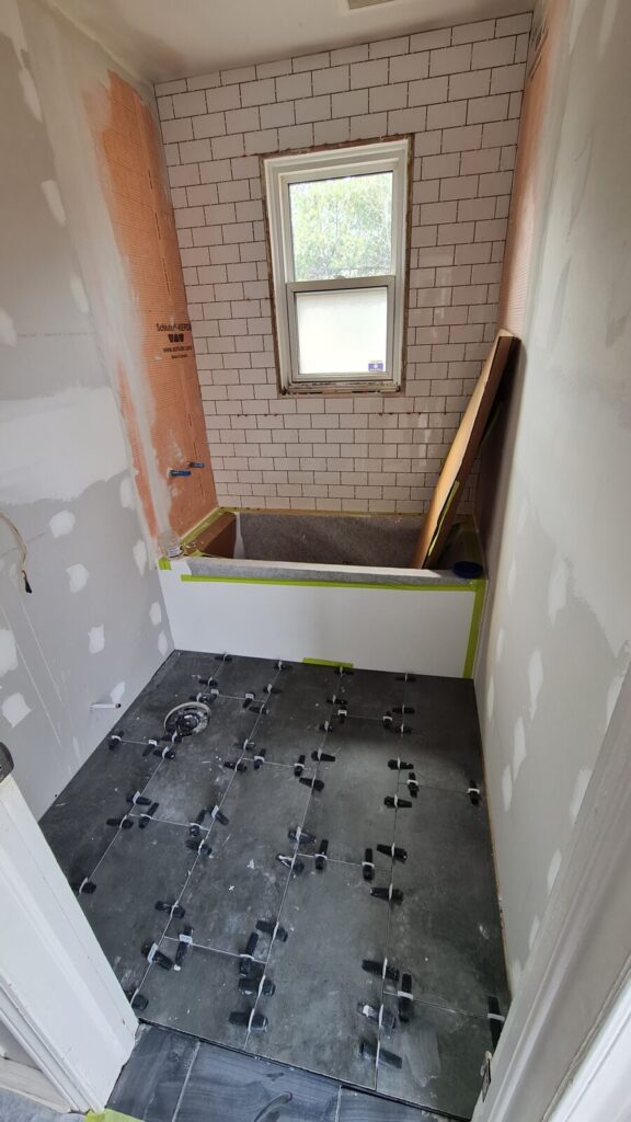 Wall and floor tiling in progress — white subway tile around the window and large-format grey floor tile being set